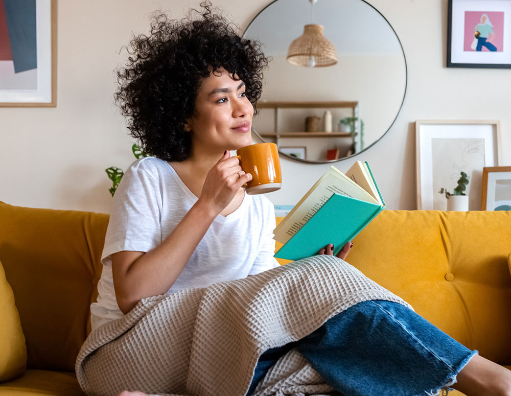 woman sitting on her couch with a mug and a book