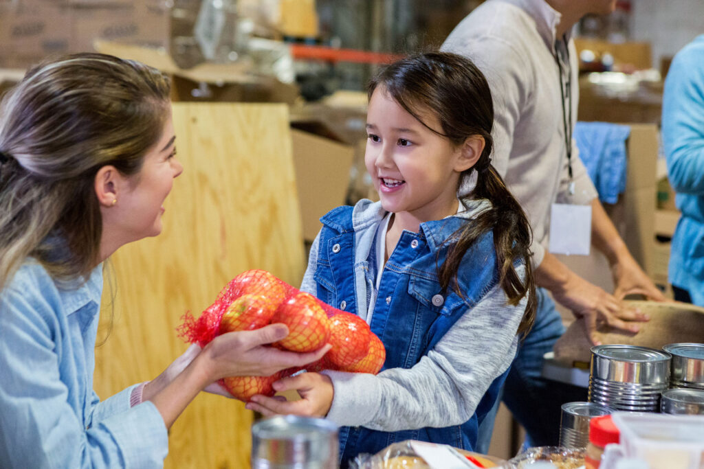Adorable little girl and her mom enjoy volunteering together in a food bank. They are holding a bag of apples.
